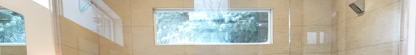 Looking through a glass shower door, through a beige tiled shower surround and out an outside window. It is a very horizontally narrow view of the shower and window.